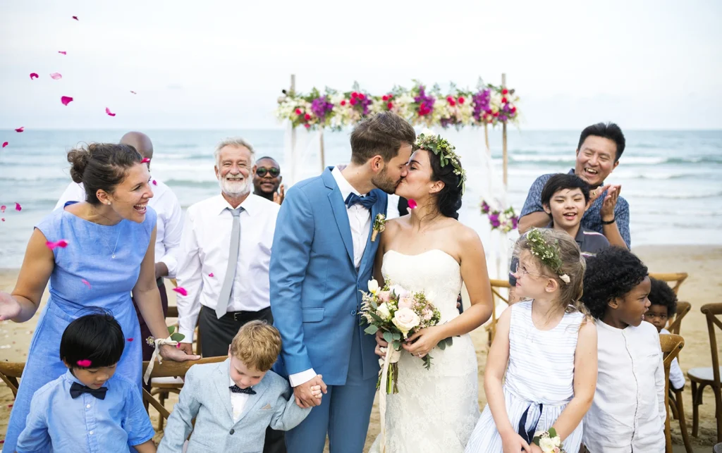 Bride and groom sharing their first kiss during a beachfront wedding ceremony in Sarasota, Florida surrounded by family and friends.