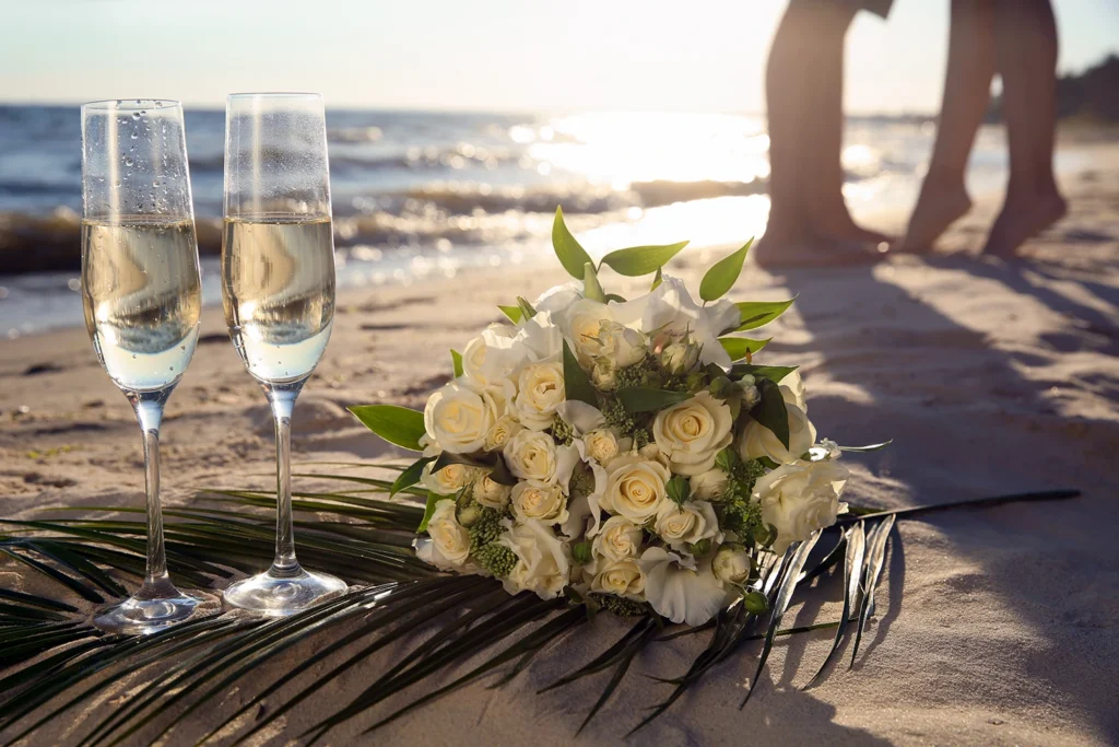Champagne glasses and bridal bouquet resting on the sand with the newly married couple walking along a Sarasota beach at sunset.